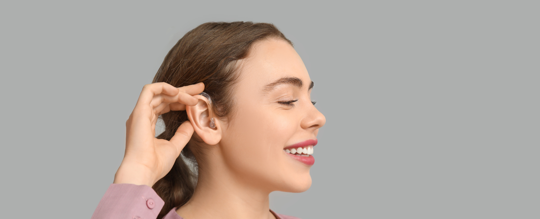 A close up of a smiling lady with brown hair and a hearing aid looking into the distance 